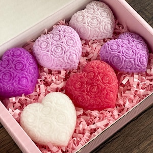 May include: A selection of heart-shaped soaps in a pink box. The soaps are in shades of purple, pink, red, and white, and feature a rose design. The box is filled with pink paper shredding.