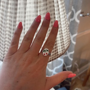 May include: A silver ring with a white oval cherry design. The ring features a pair of red cherries with green leaves. The person's nails are painted a coral color.