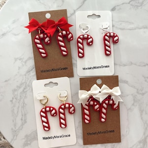 May include: Four pairs of Christmas-themed earrings. Each pair features a red and white candy cane design. Two pairs have red bows, and two have white bows. The earrings are displayed on cards with the text "MadebyMaraGrace".