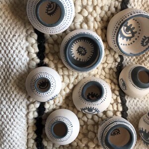 Può includere: Una collezione di piccoli vasi in ceramica bianca con disegni di occhi neri e motivi a punti neri. I vasi sono disposti su una superficie bianca e soffice.