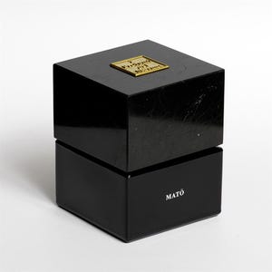 Peut inclure: Une boîte carrée noire avec une finition brillante et un couvercle. Le dessus du couvercle présente une petite plaque carrée de couleur dorée avec du texte en relief. La face avant de la base affiche le texte blanc MATŌ en police sans empattement.