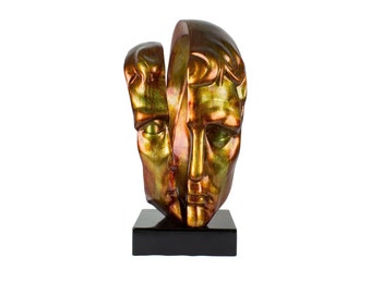 May include: Gold and green abstract sculpture of two faces, one partially obscured by the other, on a black base.