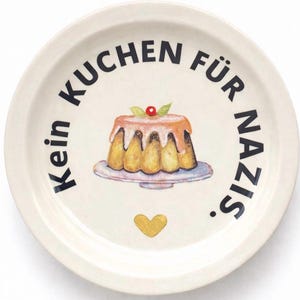 Könnte beinhalten: Eine cremefarbene Keramikplatte mit dem schwarzen Text "Kein Kuchen Für Nazis". Die Platte zeigt eine Kuchenillustration mit Kirsche und goldenem Herz.