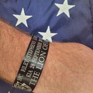 Puede incluir: Pulsera negra con texto blanco que dice "SPC RILEY STEPHENS KIA 28 SEPTIEMBRE 2004 THE LION OF"
