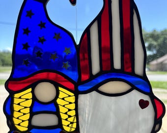 May include: Stained glass gnome couple with patriotic colors. One gnome has a blue hat with gold stars, the other has a red and white striped hat. A heart shape is formed between the two hats. A red heart is on the right gnome's cheek.