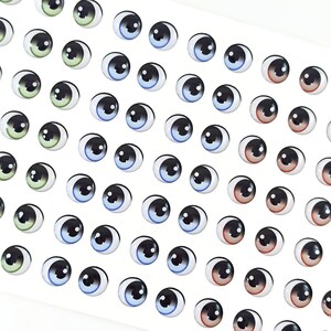 K&ouml;nnte beinhalten: Eine Blatt mit bunten Glitz Augen, in verschiedenen Farben wie Blau, Gr&uuml;n, Braun und Schwarz. Die Augen sind in Reihen auf einem wei&szlig;en Hintergrund angeordnet.