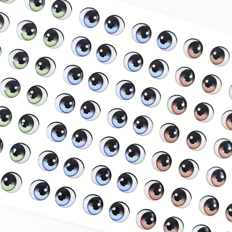K&ouml;nnte beinhalten: Eine Blatt mit bunten Glitz Augen, in verschiedenen Farben wie Blau, Gr&uuml;n, Braun und Schwarz. Die Augen sind in Reihen auf einem wei&szlig;en Hintergrund angeordnet.