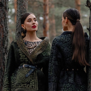May include: Two women wearing green and black quilted jackets with leopard print skirts stand in a forest. The jackets have a belt and a unique quilted pattern.