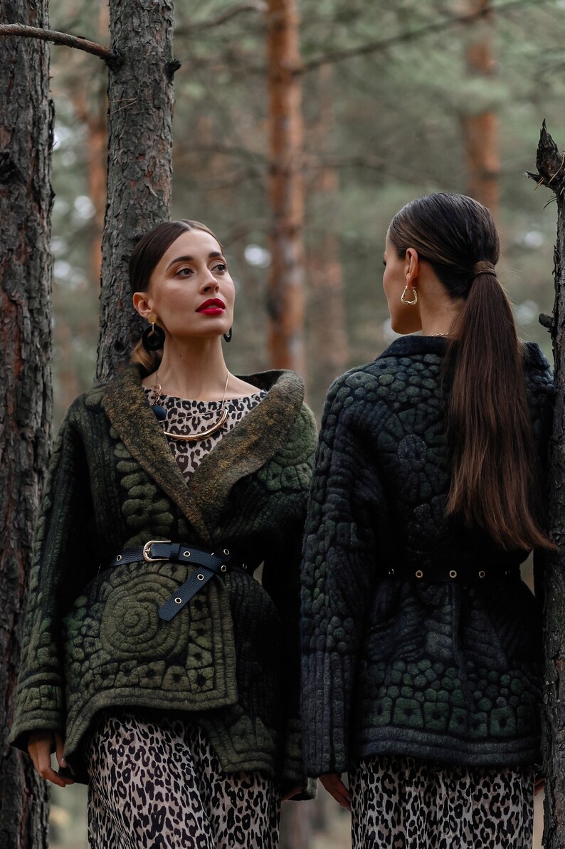 May include: Two women wearing green and black quilted jackets with leopard print skirts stand in a forest. The jackets have a belt and a unique quilted pattern.