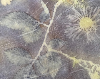 May include: A textile fabric featuring a botanical print with light yellow and cream-colored floral and leaf silhouettes set against a dark, muted purple and gray background. The pattern displays detailed impressions of various plants and flowers.