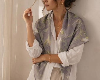 May include: A square scarf in a muted lavender-gray hue featuring a botanical print of white daisies and green foliage. The scarf is draped over a white button-down shirt and beige trousers, showcasing a smooth, lightweight fabric texture.