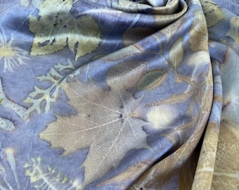 May include: A lightweight fabric scarf featuring a botanical print with various leaf and plant silhouettes in shades of tan and brown against a soft periwinkle blue background. The material has a smooth, draped texture with visible folds.