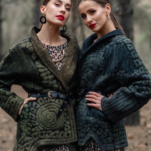 May include: Two women wearing green quilted jackets with a geometric pattern. The jackets have a black belt and a shawl collar. The women are standing in a forest setting.