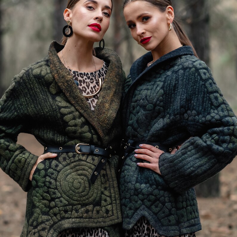 May include: Two women wearing green quilted jackets with a geometric pattern. The jackets have a black belt and a shawl collar. The women are standing in a forest setting.