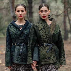 May include: Two people wearing green quilted jackets with intricate patterns. The jackets have black belts and are worn over leopard print dresses. The people are standing in a forest setting.