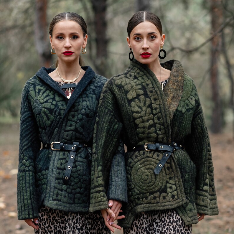 May include: Two people wearing green quilted jackets with intricate patterns. The jackets have black belts and are worn over leopard print dresses. The people are standing in a forest setting.