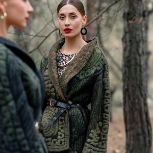 May include: A woman wearing a green quilted jacket with a black belt and a leopard print dress. She is wearing gold earrings and a necklace with a large, round pendant.