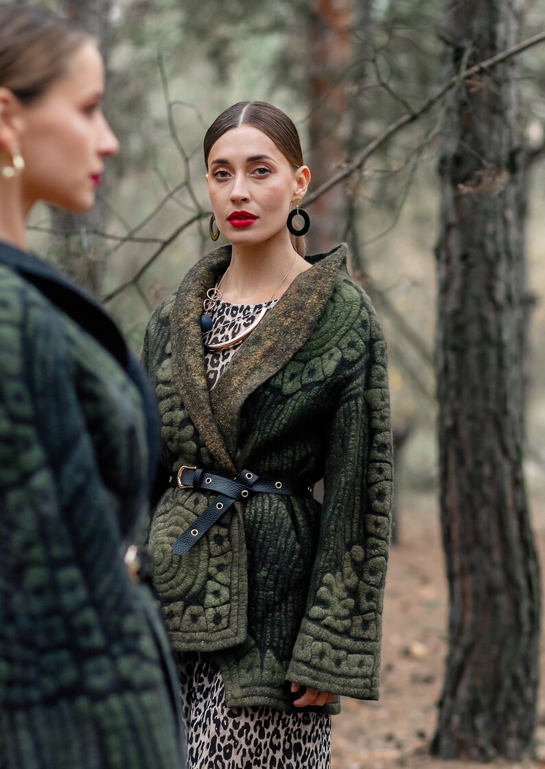 May include: A woman wearing a green quilted jacket with a black belt and a leopard print dress. She is wearing gold earrings and a necklace with a large, round pendant.