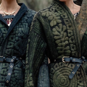 May include: Two women wearing dark green patterned jackets with black leather belts. The jackets have a unique textured design and are cinched at the waist with belts.