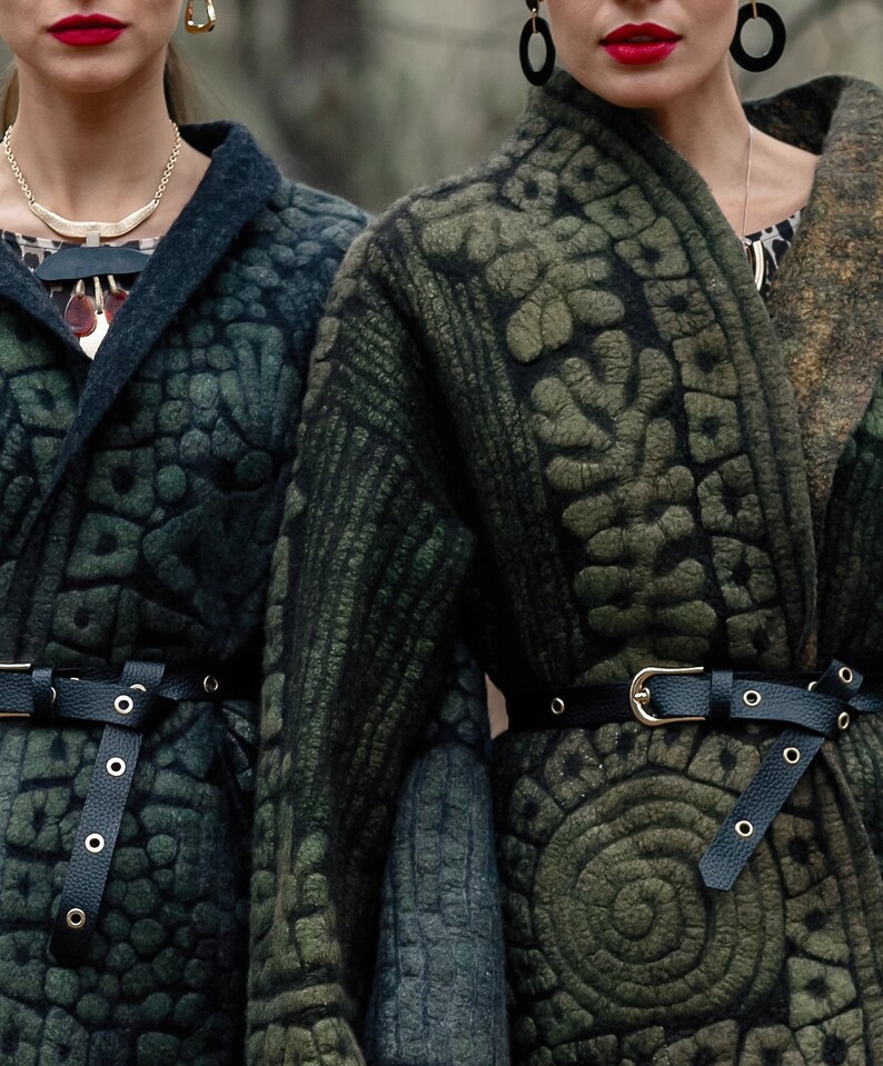 May include: Two women wearing dark green patterned jackets with black leather belts. The jackets have a unique textured design and are cinched at the waist with belts.