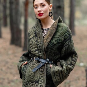 May include: A woman wearing a green quilted jacket with a black belt and a leopard print dress. She is standing in a forest.