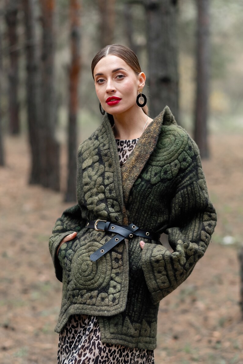 May include: A woman wearing a green quilted jacket with a black belt and a leopard print dress. She is standing in a forest.