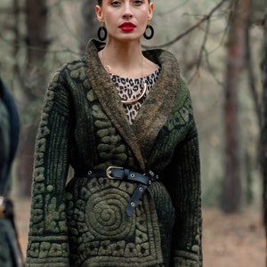 May include: A woman wearing a green patterned jacket with a black belt, a leopard print top, and gold jewelry. She is standing in a forest.