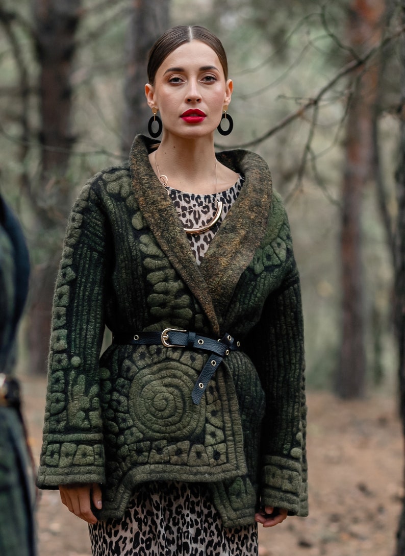 May include: A woman wearing a green patterned jacket with a black belt, a leopard print top, and gold jewelry. She is standing in a forest.