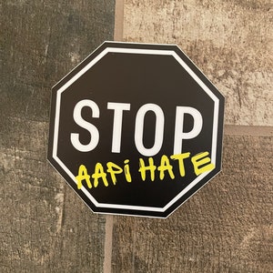 May include: A black octagon sticker with a white border and the word "STOP" in white capital letters. Below the word "STOP" is the text "AAPI HATE" in yellow, handwritten-style letters.