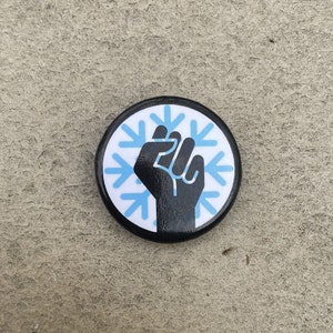 May include: A round black button with a white background featuring a black fist and light blue snowflake design. The button is a wearable accessory, likely a pin or badge, with a simple, bold graphic.