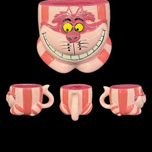 May include: A set of Alice in Wonderland-themed mugs. The large mug is shaped like the Cheshire Cat's face, with pink and white stripes. Three smaller mugs feature the same design, with handles.
