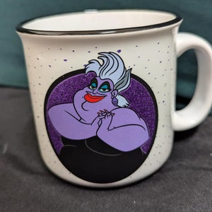 May include: White ceramic mug with a black rim and a purple and black illustration of Ursula from The Little Mermaid. The illustration shows Ursula with her signature purple skin and hair, and she is holding her hands together in front of her chest.