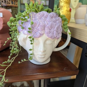 May include: A white ceramic planter shaped like a classical female bust, adorned with a crown of lavender-colored flowers. The planter holds a trailing succulent plant with green, bead-like leaves. The planter has a handle and is displayed on a dark wooden shelf.