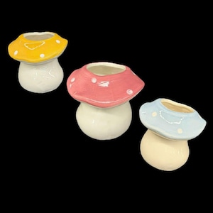 May include: Three ceramic mushroom-shaped planters. Each planter has a white stem and a colorful cap. The caps are yellow, pink, and blue, each with white polka dots. The planters are set against a black background.