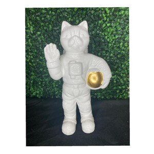 May include: White ceramic astronaut figurine of a cat with a gold helmet. The cat is wearing a white spacesuit and has its right paw raised in a wave.