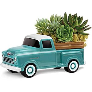 May include: A teal ceramic vintage pickup truck planter with a white roof and wheels. The truck bed holds a variety of green succulents and moss. The truck has a chrome grill and bumper.