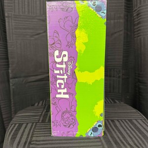 Puede incluir: Una caja rectangular con la palabra "Stitch" en blanco y el logotipo de Disney. La caja tiene un fondo morado con ilustraciones del personaje Stitch y una secci&oacute;n verde. La caja est&aacute; sobre una superficie estampada.