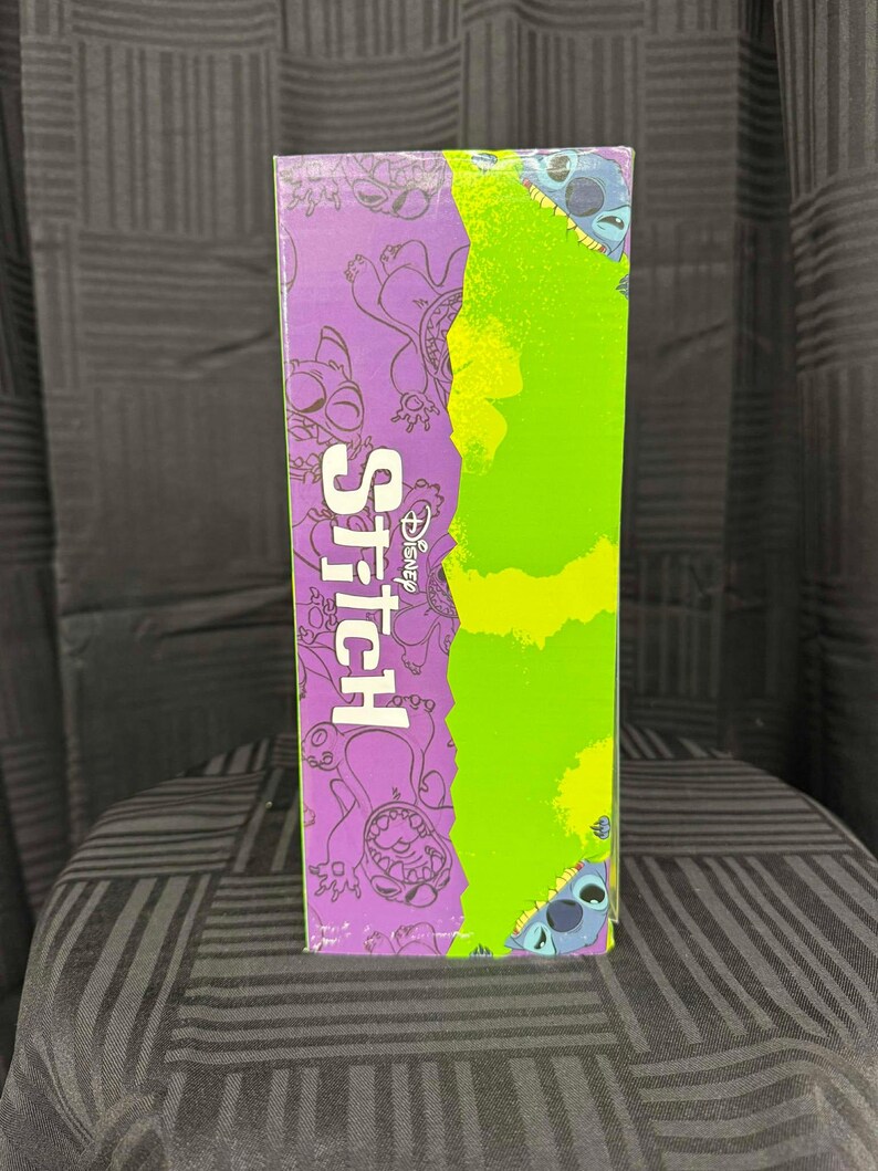 Puede incluir: Una caja rectangular con la palabra "Stitch" en blanco y el logotipo de Disney. La caja tiene un fondo morado con ilustraciones del personaje Stitch y una secci&oacute;n verde. La caja est&aacute; sobre una superficie estampada.