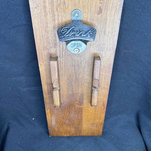 May include: A wooden wall-mounted bottle opener with a metal opener that says "Drinks" and "Open".