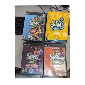 May include: Four PC game cases for The Sims 2, including the base game and the expansion packs Pets, Vacation, Nightlife, and Open for Business. The cases are all black with colorful artwork featuring characters and scenes from the game.