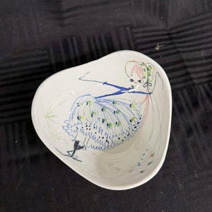 May include: White ceramic bowl with a whimsical illustration of a figure in a blue dress. The bowl has a unique, asymmetrical shape and is decorated with colorful, hand-drawn details. The artwork includes a figure with a pink hat and a blue dress.