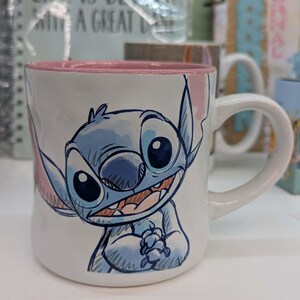 May include: White ceramic mug featuring a blue cartoon character with large ears and a mischievous grin. The interior rim is pink, and the handle is white. The mug is part of a Disney-themed collection.