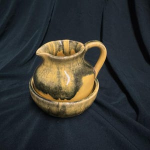 May include: A yellow and black ceramic pitcher with a wide base and a handle. The pitcher has a mottled glaze with streaks of black and yellow.