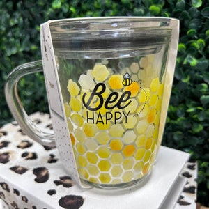 May include: A clear glass mug with a lid and handle. The mug has a honeycomb pattern with yellow and orange accents. The text "Bee Happy" is printed on the mug with a small bee illustration.