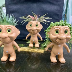 May include: Three troll-shaped planters with green eyes and a smiling face. Each planter is made of brown plastic and has a different type of plant growing out of the top. The plants are a succulent, an air plant, and a string of pearls.