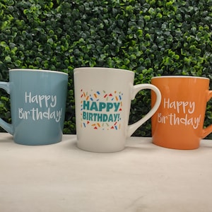 May include: Three ceramic mugs in blue, white, and orange, each with a curved handle. The blue and orange mugs have "Happy Birthday!" printed in white. The white mug has "Happy Birthday!" with colorful confetti.