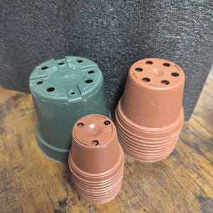 Puede incluir: Tres macetas de plástico de diferentes tamaños. Una es verde oscuro y dos son marrones. Todas tienen agujeros de drenaje en la parte inferior. Las macetas están apiladas sobre una superficie de madera.