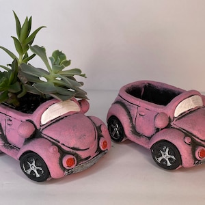 May include: Two pink ceramic Volkswagen Beetle planters. One planter is filled with succulents.