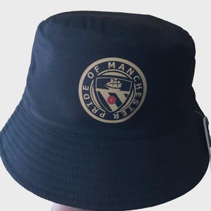 Op de afbeelding: Zwarte vissershoed met een cirkelvormig embleem met de tekst "Pride of Manchester". Het embleem toont een schip, een schild en een rode roos. De hoed is gemaakt van een zacht materiaal en heeft een brede rand.