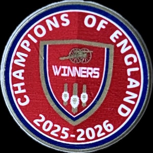 May include: A circular enamel pin in red, white, and blue, with the text "CHAMPIONS OF ENGLAND". The center features a shield with "WINNERS" and the years "2025-2026". The pin has a black background.